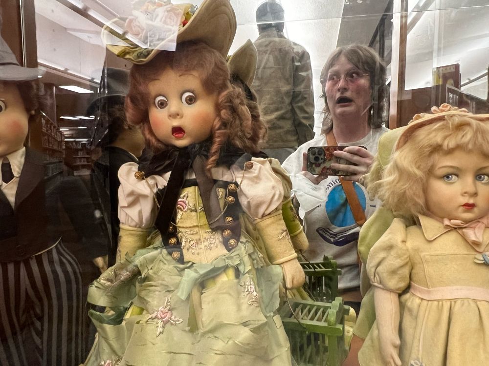 A display of antique dolls, one of whom is wearing Victorian style dress and making a terrified facial expression. To her right is a blonde doll wearing 1920s dress with an expression that says “I’m over it.” Reflected in the mirrors back of the display is me, Beth, making a matching terrified facial expression and holding my phone. 