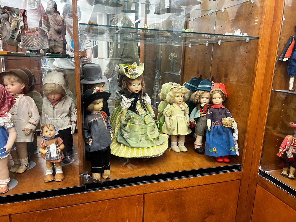 A selection of around a dozen porcelain dolls in a mirror backed display case. The dolls have a range of clothing and hair styles with variance in era (Victorian dress for example) and culture (a boy in lederhosen, for example), but not in ethnicity. All of the dolls are white.