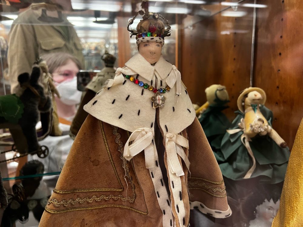 Front and center is a doll wearing King’s attire, including a crown and cloak. His expression is serene. To his right is a cow figurine wearing a dress with a hat, and is holding a floral bouquet. She’s gently smiling. 