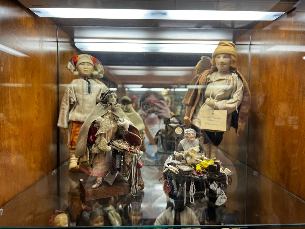This display case has four dolls. 

The smallest is about four inches tall. She is an old woman with a porcelain face sitting at a table covered in miscellaneous house goods from the early 1900s, such as tied scrolls, a handbag, a small milk pitcher, candles, and more. 

To her left is a figure about six inches tall on a one inch wooden platform. Her face is made of a very finely knit fabric. She is an old knit goods sales woman wearing many layers of cold weather clothing including a cloak, and she is holding a bag of her wares. 

Behind her is a taller figurine, around ten inches, who is made of porcelain. She is wearing what I believe to be traditional Ukrainian clothing including a jaunty hat and fuzzy earrings.

Finally, the tallest figurine is around a foot tall. His face and hands are very finely knit, and because he was purchased around a hundred years ago he is wearing a tag reading “Russian peasant from Ukraine.” He has blue eyes and a sweet smile. His blonde hair is mostly hidden by a golden yellow hat. 


