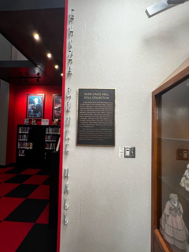 Right side of the photo. 
A white wall has a plaque reading: ELIZA CRUCE HALL DOLL COLLECTION.
ELIZA CRUCE HALL GAVE HER PRIZED DOLL COLLECTION TO THE ARDMORE PUBLIC LIBRARY IN 1971. SHE HAD SPENT THIRTY FIVE YEARS COLLECTING DOLLS FROM AROUND THE WORLD AND AS SHE APPROACHED HER EIGHTIETH BIRTHDAY, SHE DECIDED IT WAS TIME TO PROVIDE FOR THEIR FUTURE, SHE WANTED CHILDREN TO ENJOY HER DOLLS AS THEY HAD ON VISITS TO HER HOME, AND SHE CHOSE THE LIBRARY AS A PERFECT NEW HOME FOR HER CHERISHED COLLECTION. THREE WEEKS AFTER THE DEDICATION OF THE COLLECTION, MRS HALL DIED DURING AN AFTERNOON NAP.
MRS, HALL HAD LIVED IN ARDMORE SINCE HER FATHER BROUGHT THE FAMILY HERE FROM MARION: KENTUCKY IN 1896, HER FAMILY WAS INFLUENTIAL IN THE DEVELOPMENT OF ARDMORE AND THE STATE, HER UNCLE LEE CRUCE WAS THE SECOND GOVERNOR OF OKLAHOMA.

On the left side of the photo is a twilight themed reading nook, with red and black checkered carpet and red walls.