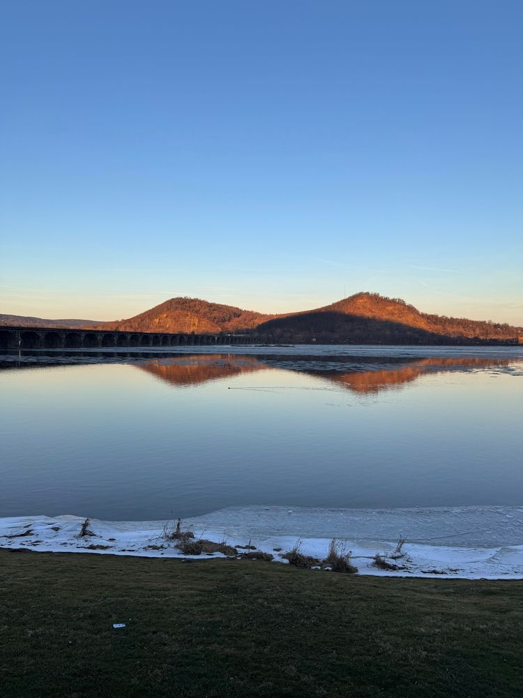 This image, taken from the Bridgeview Bed and Breakfast, showcases a tranquil scene featuring the Susquehanna River in the foreground. The historic Rockville Bridge spans the river, and in the background, Blue Mountain rises prominently, partially illuminated by the warm light of sunset. The clear sky enhances the peaceful atmosphere. Also there’s a mallard duck in the water. 
