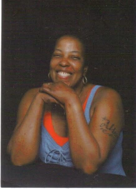 My mother. This photoshoot was about 6 weeks before she passed away. 
She’s a medium brown skinned Black woman of indeterminate age due to being fully Black lol. Nah she looks about 35 in the pic but she’s 44. She’s wearing a blue denim strap dress with a red (her fav color) tank top underneath. You can see the first 3 letters of her only tattoo “Ali-“ which is for Alice. 
She has a wide, full, cheeky smile. Her hands are resting on her chin. 
It’s been 17 years since this pic was taken. I miss her dearly. 