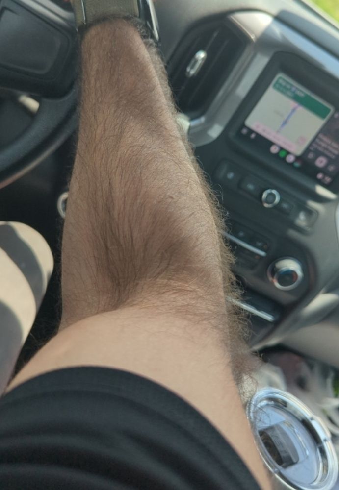 A man's arm and hand holding a steering wheel from inside the cab of a vehicle, a grassy field is visible outside the windshield