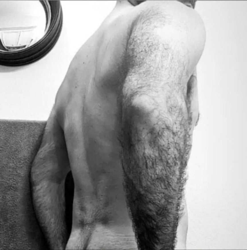 A black and white side/rear profile of a man's back and hairy arms standing in a white room, a small circular mirror hanging on the wall to the left