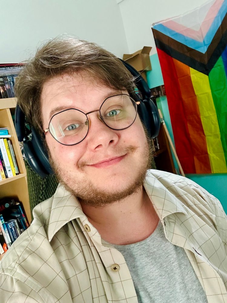 A picture of me wearing headphones and at my desk, vest and overshirt in my office and behind me is the LGBT flag. I’m leaning back a lot, not quite face on.