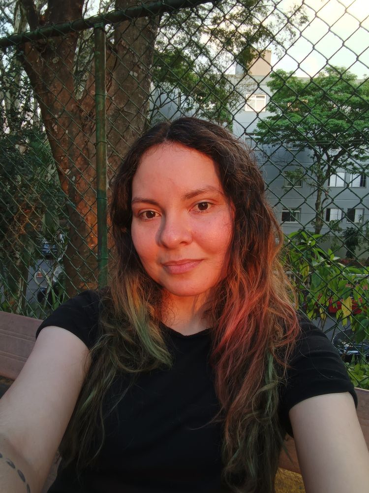 Foto minha de camiseta preta, cabelo ondulado cacheado castanho em cima, verde e rosa embaixo. O sol está no golden hour