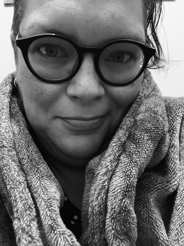 A black and white image of a woman wearing glasses and wrapped in a fluffy dressing gown. She has a wry smile. 