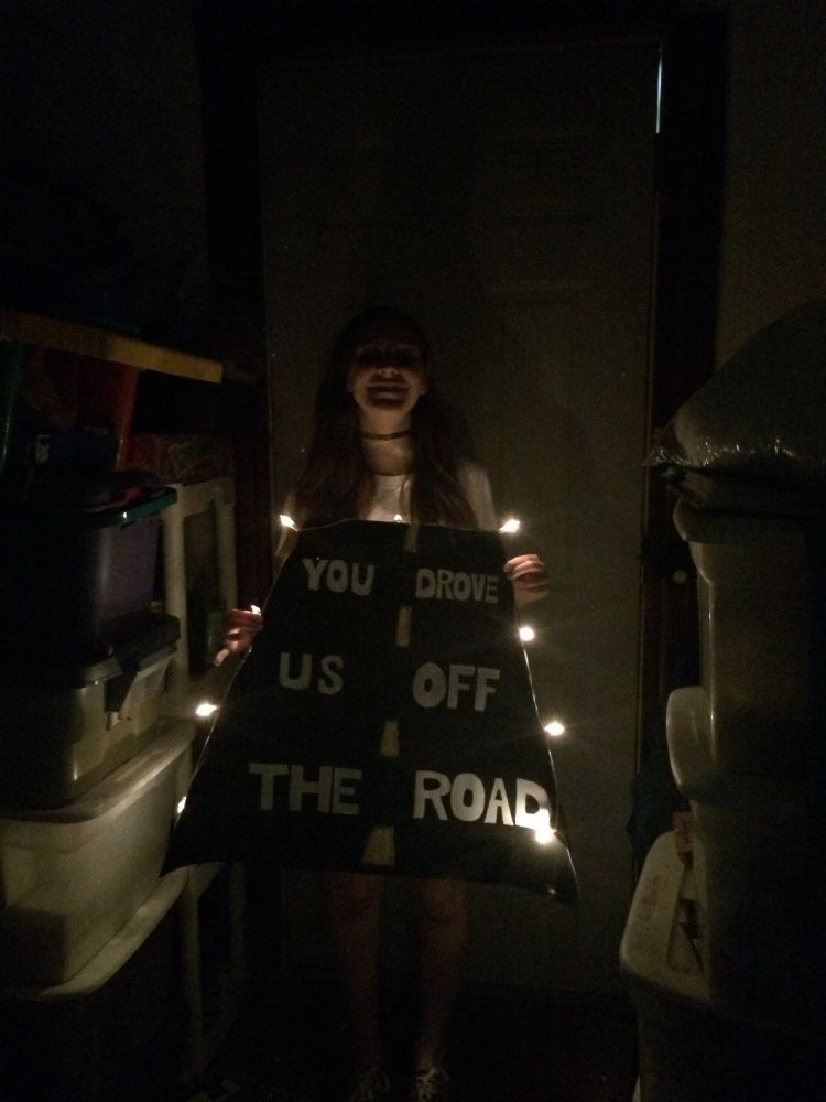 cynthia in a dark room holding a black sign with lights around it. the sign says "you drove us off the road".
