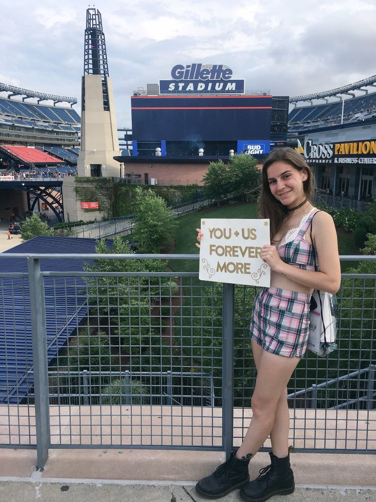 cynthia on the overlook at gillette stadium holding a sign that says "you + us forever" in gold letter stickers. i am wearing a two peace teal and pink plaid set, with doc martens. 
