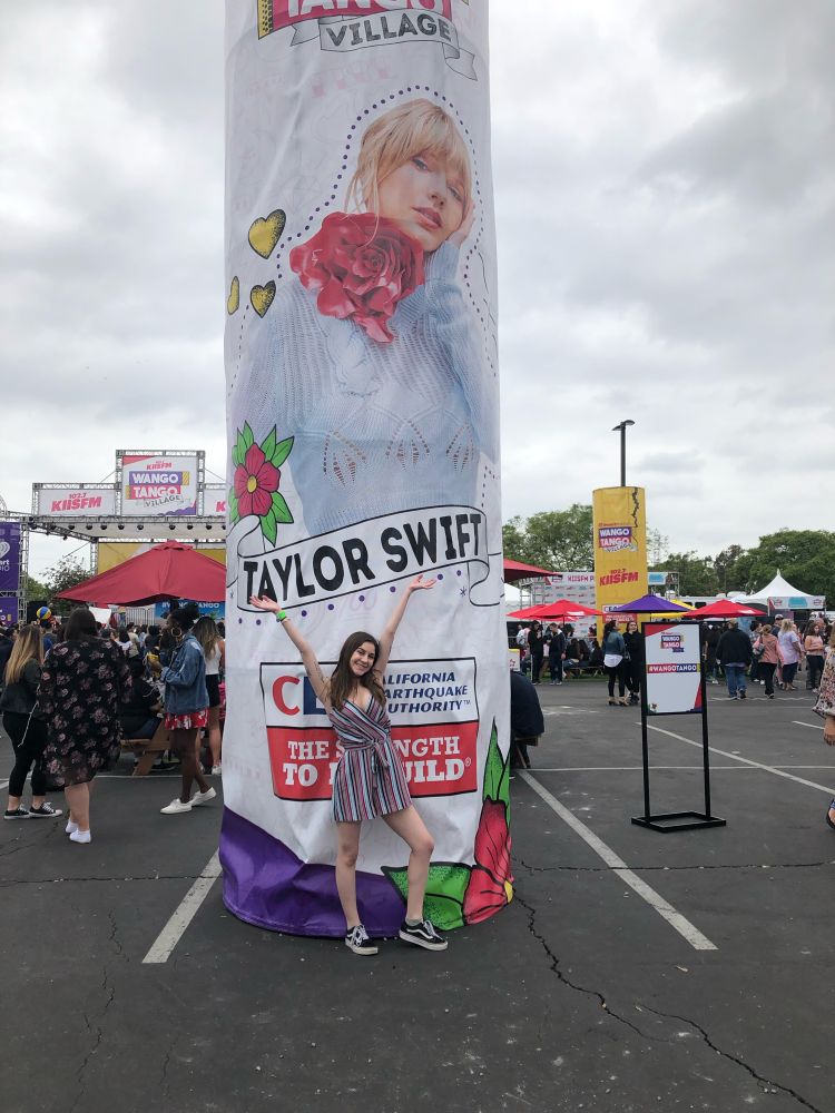 cynthia at wango tango in front of a giant picture of taylor swift from the lover promo, it is set in a parking lot. i am wearing a mutli colored teal romper and vans.
