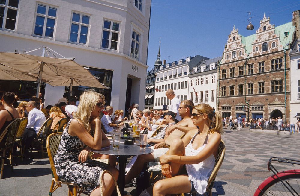 A European town square, surrounded by low, centuries-old buildings. People sit at tables enjoying the sunsnd each others company.