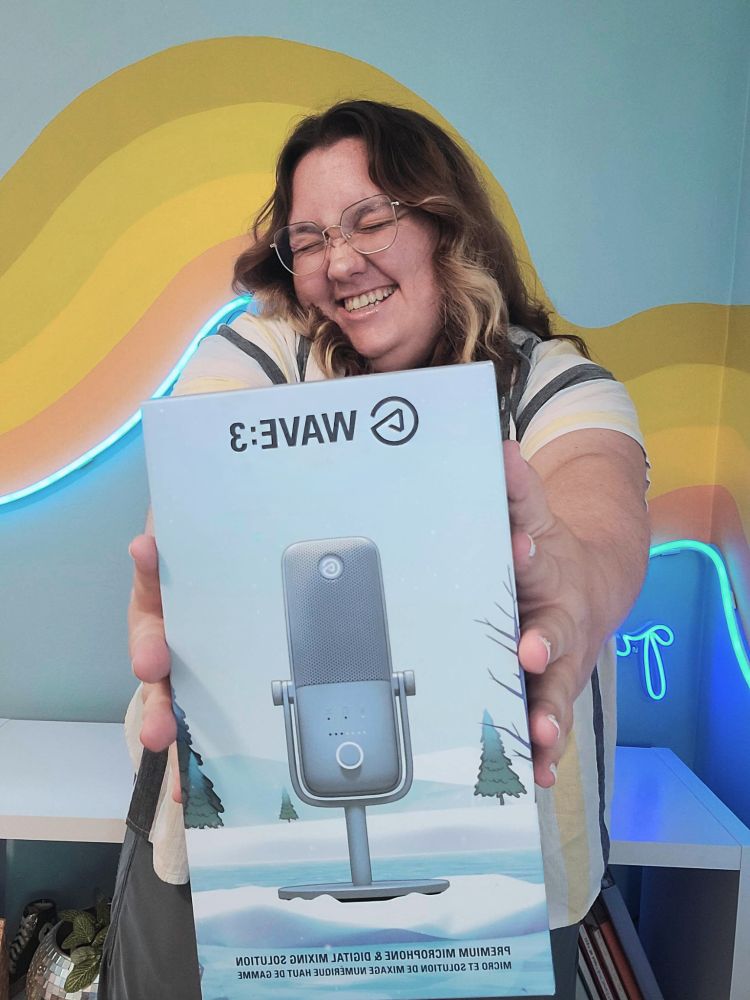 myself, a white nonbinary person who presents pretty femininely with midlength wavy brunette hair with some blonde pieces in the front from a grown-out money piece. i'm wearing glasses, a striped shirt and blue pants making a squinty smiley face as i hold out a light blue box featuring the elgato wave:3 microphone in light blue glacier freeze color with wintery background. i'm standing in front of a mural in my room with a wave effect in green and orange shades on a blue background and some blue neon lights and a white shelf. 