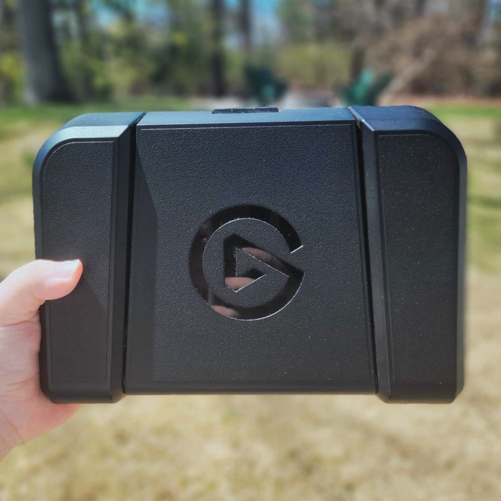 a black elgato foot pedal with the shiny elgato logo. the background is a blurred grassy background and you can see my white hand holding it on the left.