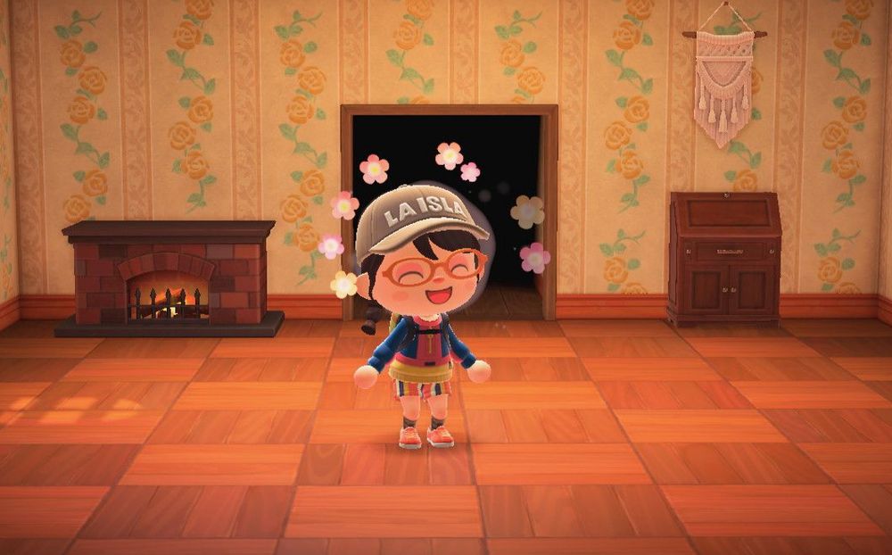 animal crossing new horizons character with brown hair that is in a braid to one side and has bangs wearing a tan hat, a multicolored jacket and pants, orange shoes and a backpack as well as orange glasses smiling with flowers around them in a room with reddish brown tone wood on the floor in a parquet style, a fireplace, and armoire as well as a cloth tapestry. the wallpaper has yellow roses and a matching wood border.