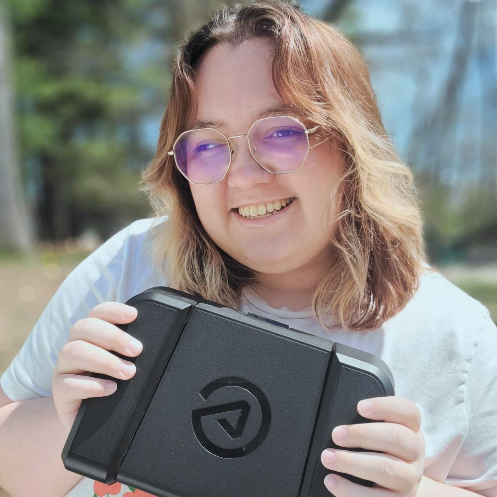 a photo of me, a white plus-size nonbinary person with shoulder length wavy brown hair with some blonde bits in the from growing out a money piece. i'm wearing wire rim glasses, a white shirt, and i'm holding a black elgato foot pedal. the background is a blurred blue sky with trees.