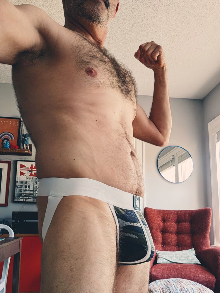 A standing pose in a camo-and-white gruffpup jockstrap. The focus is on my hairy chest and pit, and also my pronounced bulge. The photo is cropped at my scruffy chin and my upper thighs.