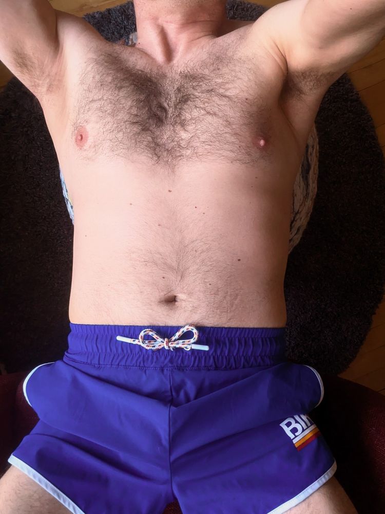 A cropped photo in which I'm wearing electric blue Bike shorts, with my arms above my head to reveal some pit and chest hair.