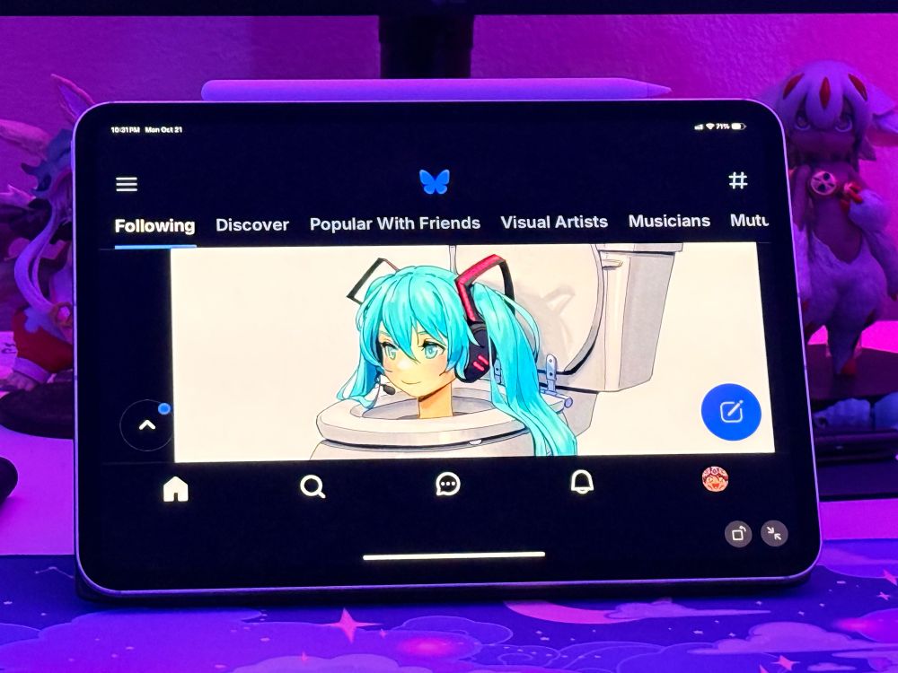 Photo of an iPad running Bluesky’s iPhone app in landscape mode. The feed is displaying a Hatsune Miku Skibidi Toilet artwork by @pikat.io. There are anime figures from “Made In Abyss” in the background, and the room lighting is purple.