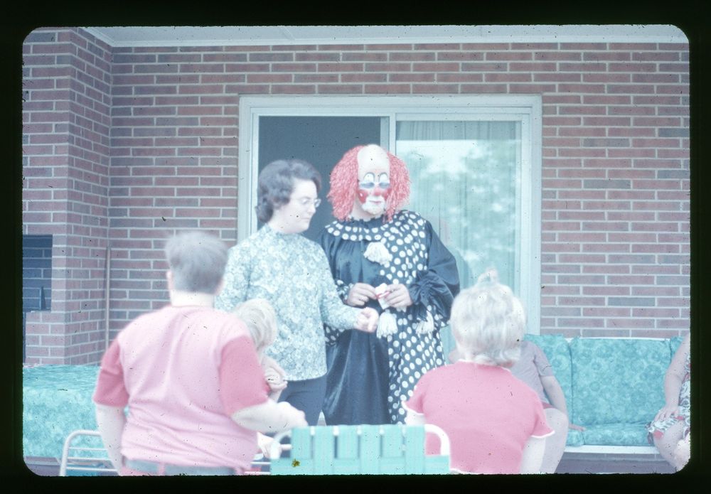 The image shows a small group of people gathered on a patio with a brick backdrop. A person in a clown costume with bright red hair and a polka-dot outfit stands alongside a woman in a patterned blouse. Seated in the foreground and background are several children and adults, some wearing casual attire. The clown has a pack of smokes. He doesn't get paid enough for this shit.