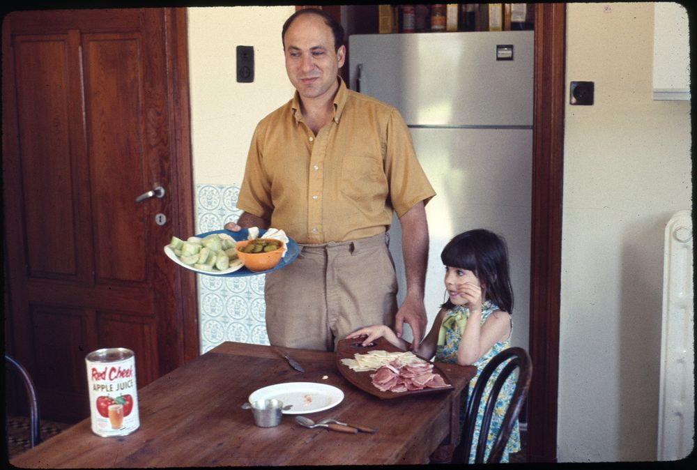An adult man stands in a kitchen holding plates with vegetables and pickles, while a young girl in a floral dress stands beside him. On the wooden table are a can of Red Cheek apple juice, a plate with sliced meats and cheese, and utensils. The room features light-colored walls, a wooden door, and a refrigerator in the background.