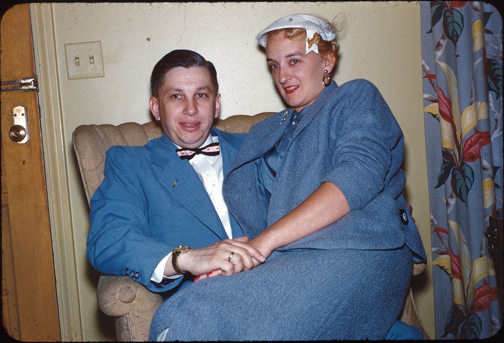 A couple dressed in blue suits is seated indoors. The man, wearing a blue suit with a bow tie, has his hands clasped with the woman sitting on him. The woman wears a matching blue suit and a white hat with a bow. Behind them is a curtain with a colorful floral pattern. The setting suggests a cozy indoor environment with a light-colored wall and a visible light switch.