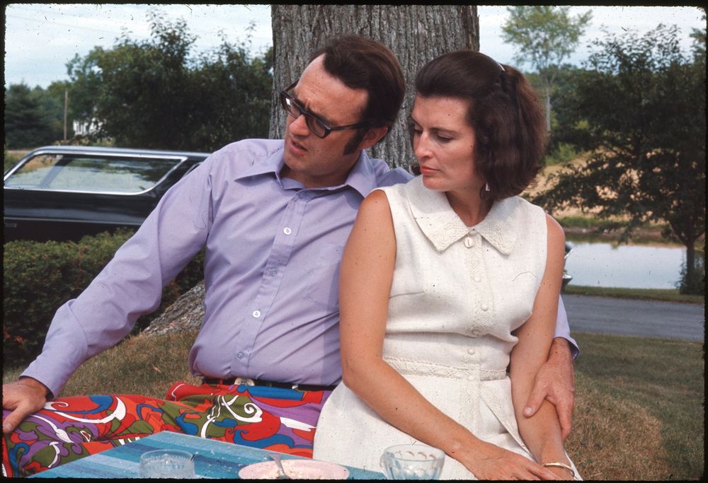 A man and woman are sitting together outdoors next to a tree. The man is wearing a purple shirt and brightly patterned pants, while the woman is dressed in a sleeveless white dress. They appear to be sitting on a grassy area near a road with a parked car visible behind them. The background includes green foliage and glimpses of a distant pond or body of water. The table in front of them has some glassware and a bowl. The pants are awesome.