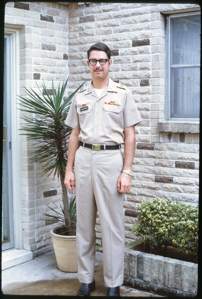 The image shows a man in a military uniform standing outside a brick building. He is wearing glasses and has short dark hair. There's a potted plant next to him and a small shrub in the foreground. The image is taken during daylight.