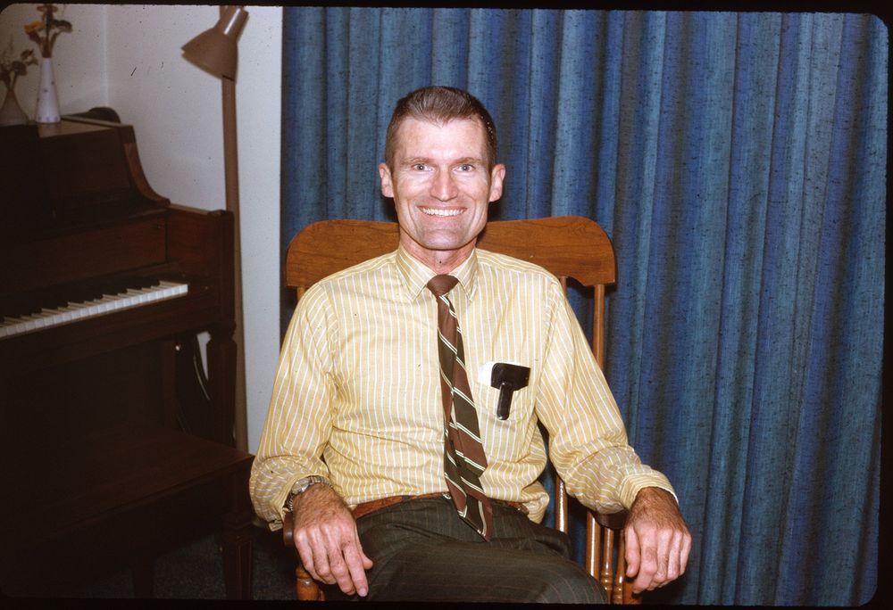 A smiling man sits in a wooden chair indoors, wearing a yellow striped shirt and a tie with a green and brown pattern. Behind him are blue curtains and a piano partially visible on the left side. A lamp and decorative vases are placed on the piano, indicating a cozy home setting.