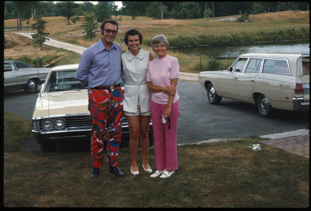 The image shows three people standing outdoors near two parked cars on a driveway. A man in colorful pants and glasses stands next to two women, one wearing a light-colored dress and the other in a pink outfit holding sunglasses. They are positioned in front of a beige sedan and a light-colored station wagon. The background features a grassy area with a pond and scattered trees. His pants are amazing.