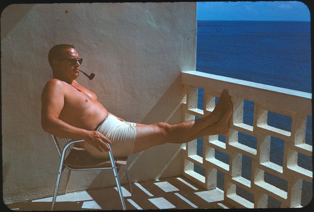 A middle-aged man sits on a balcony chair, wearing shorts and sunglasses, and smoking a pipe. His legs are propped up on a breezy concrete barrier, with the expansive ocean visible in the background under a clear blue sky.