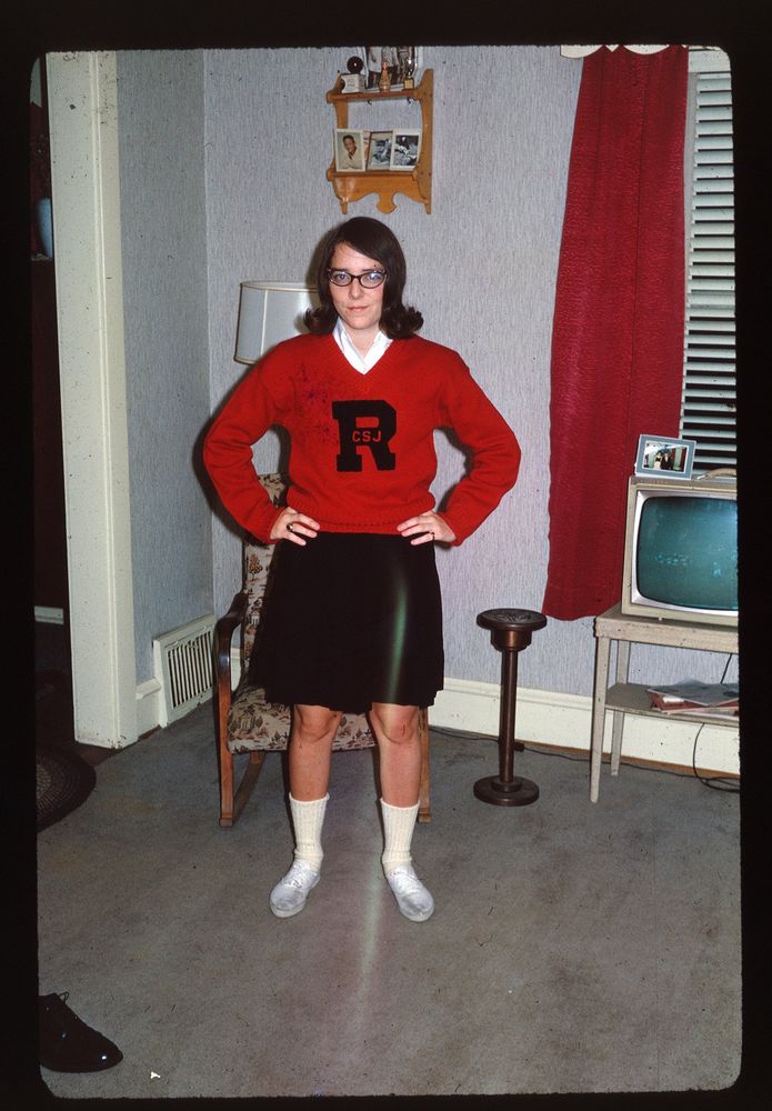 The image shows a young woman standing in a living room, wearing a cheerleader outfit: red sweater with the letter 'R' and 'CSJ' on it, a black skirt, white socks, and white shoes. She has short dark hair and glasses, with hands on hips. The room has a patterned armchair, a small television on a stand with a picture frame next to it, and a red curtain on the window. The walls are light-colored, and there is a small shelf with photos on the wall.