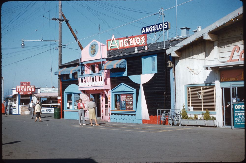 A lively image of a waterfront area featuring a colorful building labeled 'Angelo's' with a vibrant pink and black facade. Several people are walking or standing in front of the establishments, which include a restaurant and a fishing excursion service. Visible signage advertises seafood and drinks alongside fishing opportunities. Monterey, California, c. 1950s/60s.