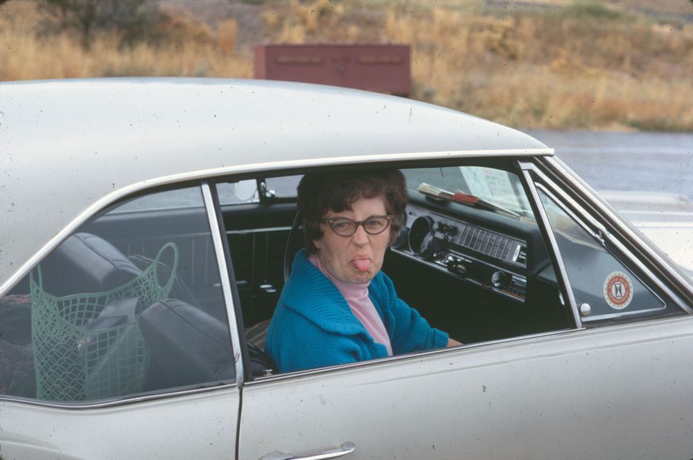 A woman wearing glasses and a blue sweater is seated in the driver's seat of a silver vintage car, sticking her tongue out playfully. The vehicle's interior shows a dashboard with classic design elements. The background includes dry grass and part of a road.