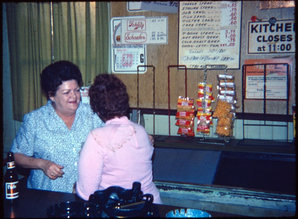 The image depicts two women conversing in a restaurant setting. One is wearing a blue patterned blouse, while the other has her back to the camera. A variety of snack items, including cornn chips and cheese puffs, are displayed alongside a menu featuring various sandwiches and steak options. A beer bottle is visible on the counter. Signs indicate the restaurant closes at 11:00 PM, and brands like Schlitz and Schaefer are advertised.