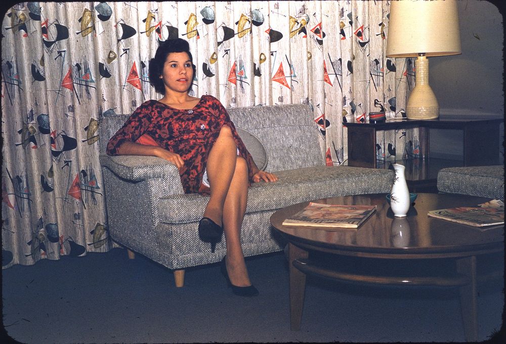 A woman sits on a textured sofa in a living room setting with distinct mid-century modern decor. She is wearing a red patterned dress and black shoes. The backdrop features curtains with an abstract geometric print, and a lamp stands on a side table with similar retro style. A round wooden coffee table holds magazines and a decorative vase.