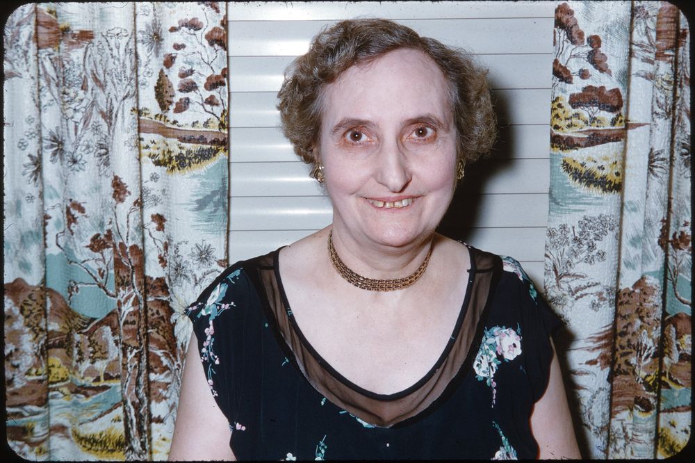 The image shows a woman with short, curly hair and a warm smile standing indoors. She is wearing a dark floral dress with sheer detailing at the neckline and a gold choker necklace. The backdrop features curtains with a landscape-themed pattern, including trees and natural scenery.