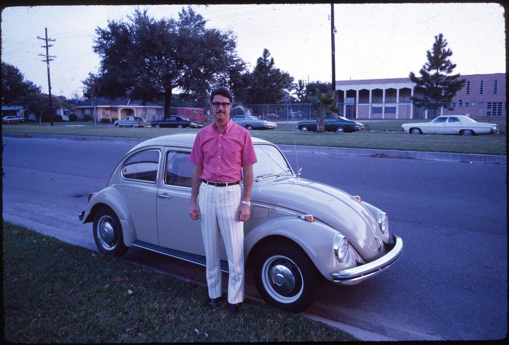 A man in a bright pink shirt and striped white pants stands beside a beige Volkswagen Beetle parked on a residential street. The background includes several parked cars, a large tree, and buildings enclosed by a chain-link fence.
