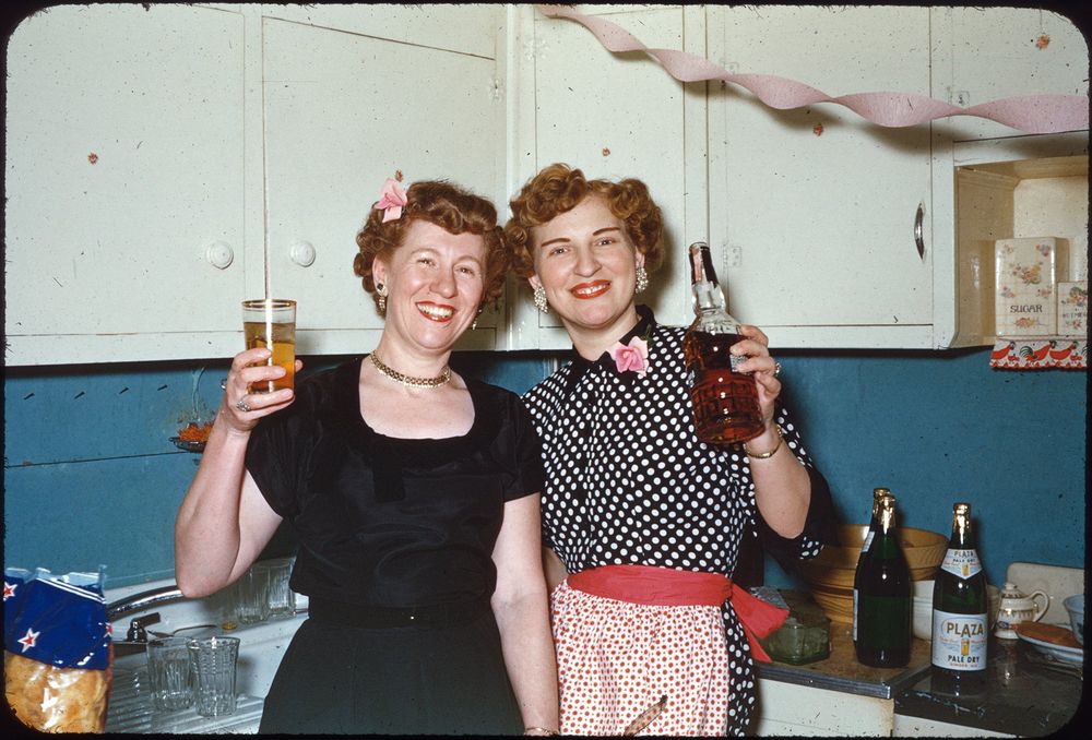 Two women are smiling and celebrating in a kitchen. One woman is holding a drink in a tall glass, and the other is holding a bottle of liquor. Both are dressed in vintage-style attire, with one in a black dress and the other in a polka dot dress with a red belt and floral accessory. The kitchen has white cabinets and a blue backsplash. Streamers hang decoratively across the room, and there are bottles and glasses on the countertop.