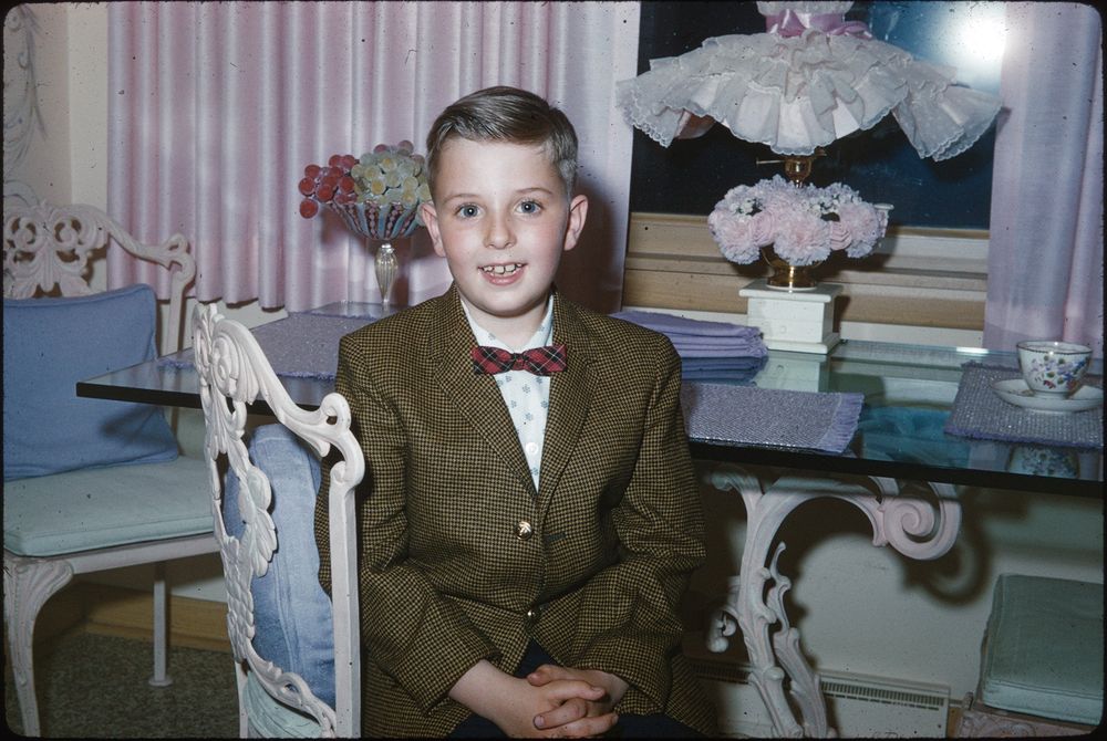 A young boy is sitting indoors, dressed in a brown checkered blazer, a white shirt with a small pattern, and a red bow tie. He is seated on an ornate, white chair at a glass-topped table. In the background, there are light pink curtains, a decorative lamp with a frilly shade, flowers, and a patterned cup and saucer on the table.