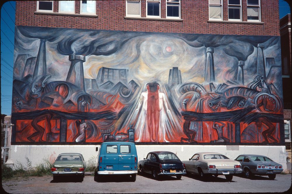 The image shows a large mural painted on the side of a brick building. The mural depicts an industrial scene with smokestacks and abstract figures, incorporating vivid colors like red and gray. Below the mural, five vehicles, including a blue van and various cars of the era, are parked in a row. The scene is well-lit, suggesting clear weather conditions. The 35mm slide was dated May 16, 1977. Location is near 26th and Folsom St in San Francisco.
