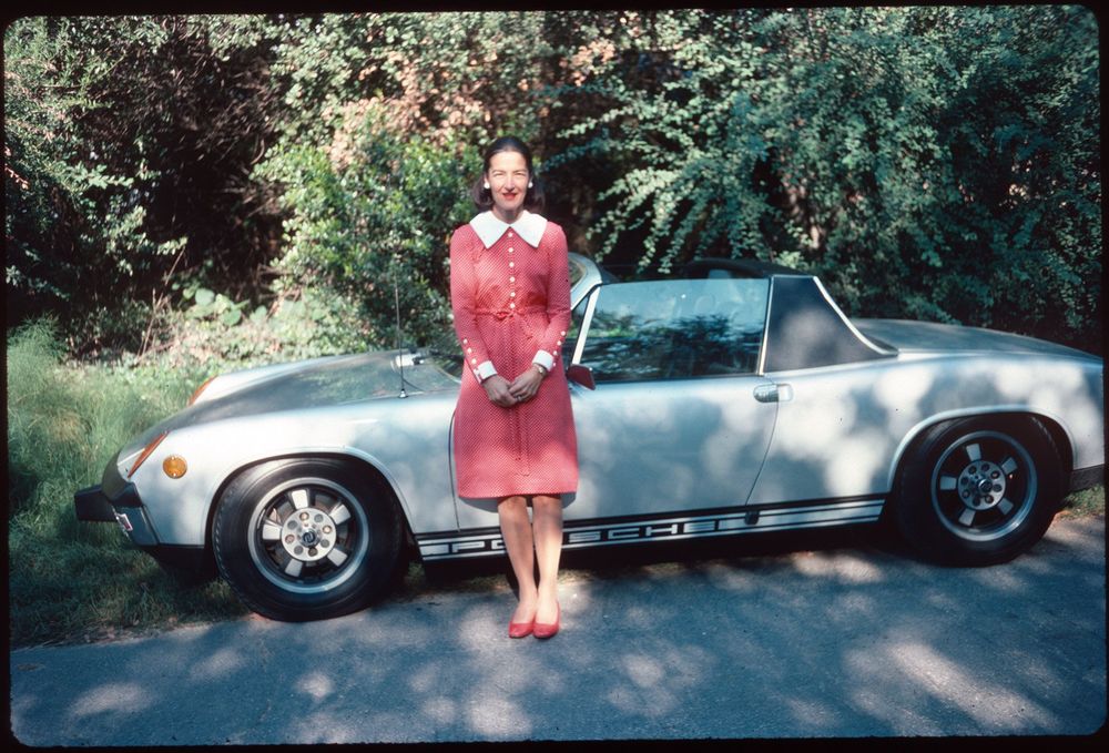 A woman in a red dress with a white collar stands in front of a silver Porsche sports car on a sunny day. The car is parked on a paved road with lush green foliage in the background.