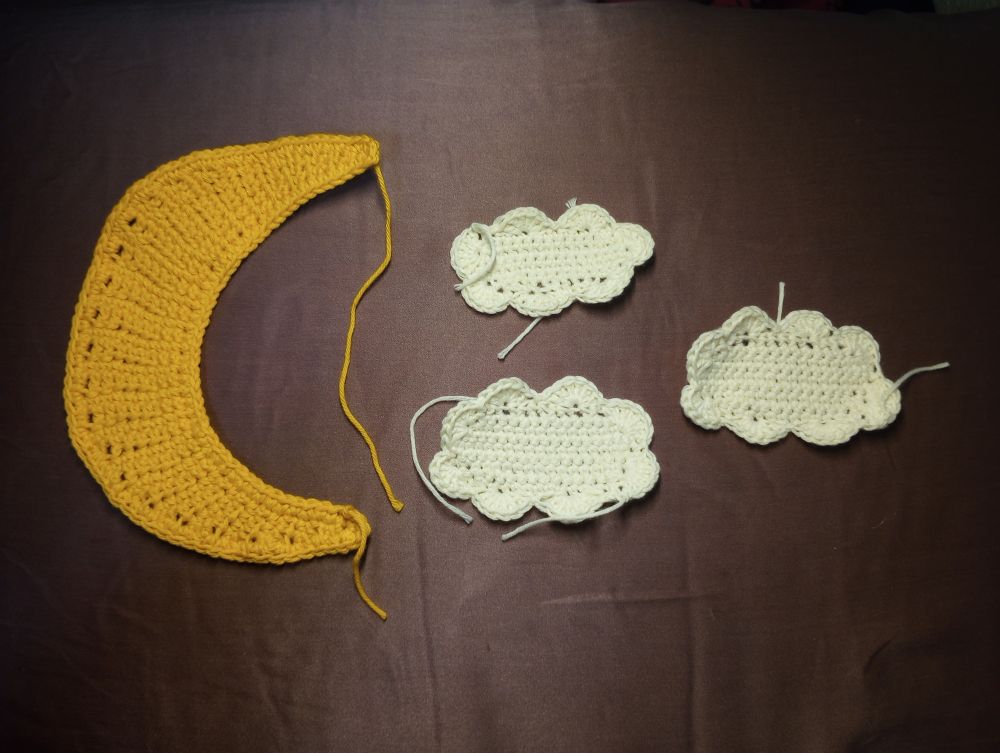 Photo of a crocheted crescent moon made with yellow yarn next to three crocheted clouds made with white yarn that are all slightly different sizes.