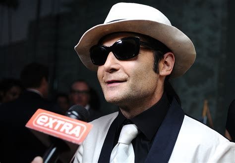 a man, actor corey Feldman, is pictured in a white hat, white jacket, and dark sunglasses talking into a microphone.