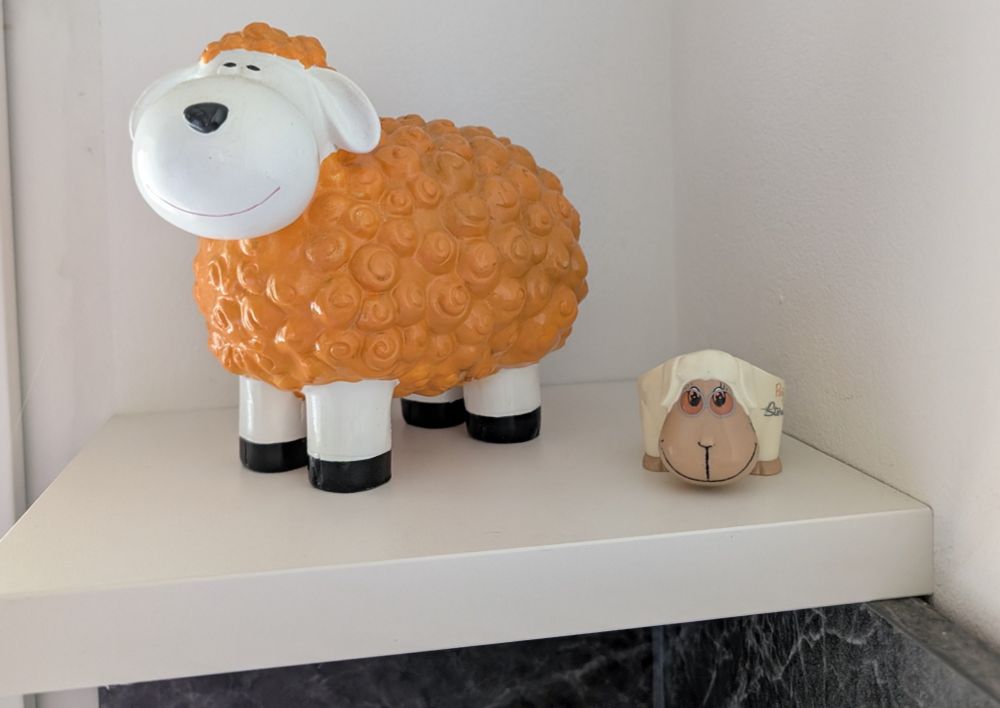 Man sieht auf ein kleines weißes Eckregal-Brett. Darunter schwarze Fliesen. Auf dem Brett steht ein weißes dickes Keramikschaf, dessen Wolle am Körper und am Kopf orange gefärbt ist. Rechts daneben ist noch ein viel kleineres Schaf, ganz in eierschal-weiß. Die Schafe wirken, als ob sie lächeln.