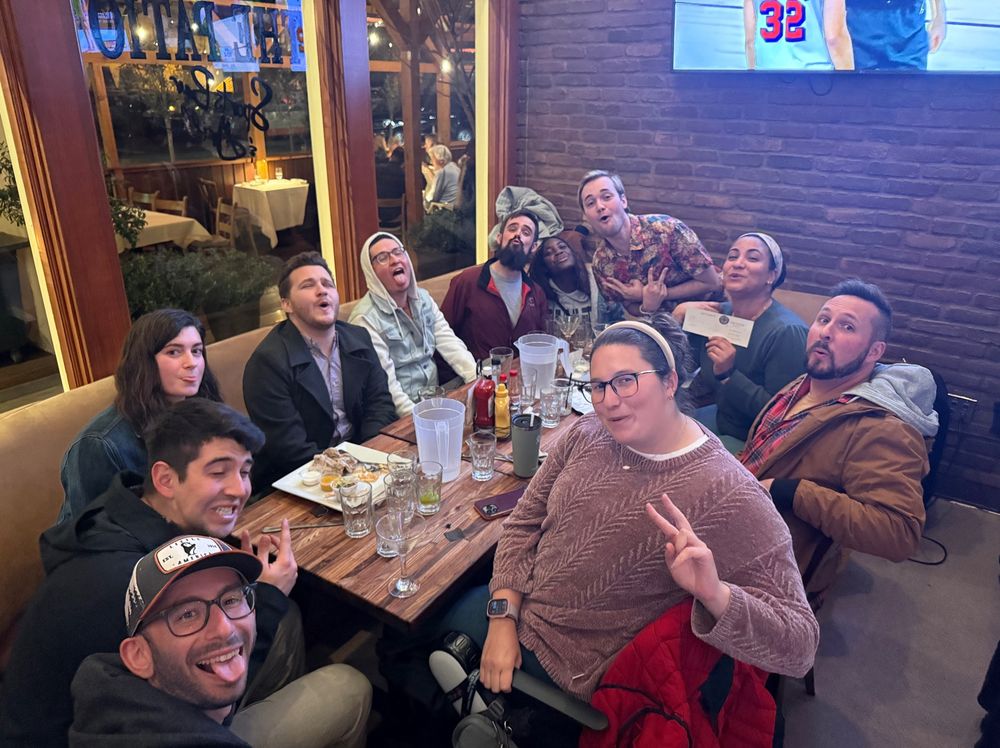 A group of 11 people sit at a table in a bar, looking at the cameras. They are smiling and howling 