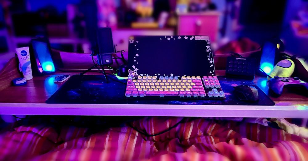 A wood-effect over bed-desk with a metal frame, with keyboard, mouse, art tablet, rgb speakers, galaxy desk mat among other things sat on top of it.