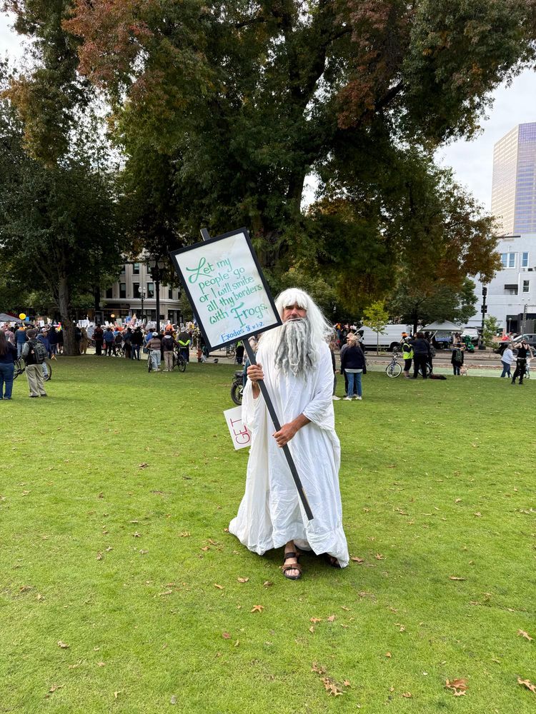 A man dressed in a white robe and a white beard that looks like God holds a sign that reads “Let my people go or I will smite thy borders with frogs. Exodus 8 v1-2”