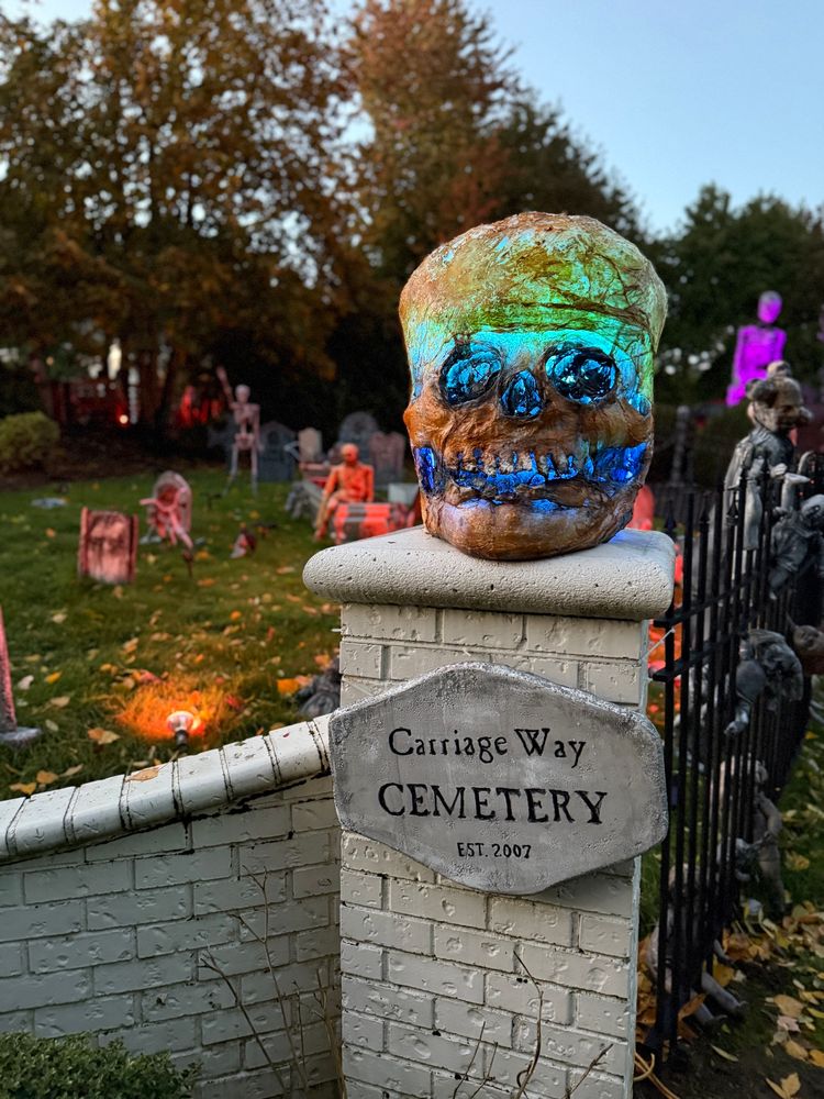 A huge skull with blue and aquamarine light coming out of it sits atop a brick fence post with a sign that reads Carriage Way Cemetery est 2007. 