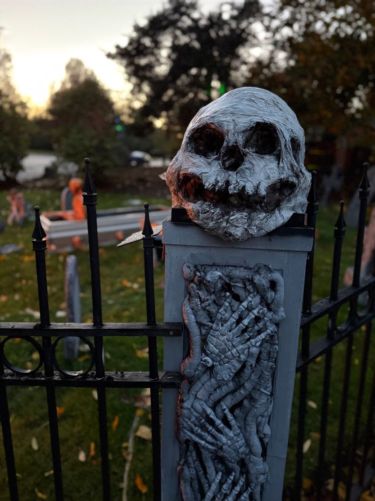 An enormous distorted skull rats on a pillar recorded with skeletal hands that joins two sector wrought iron fence. 